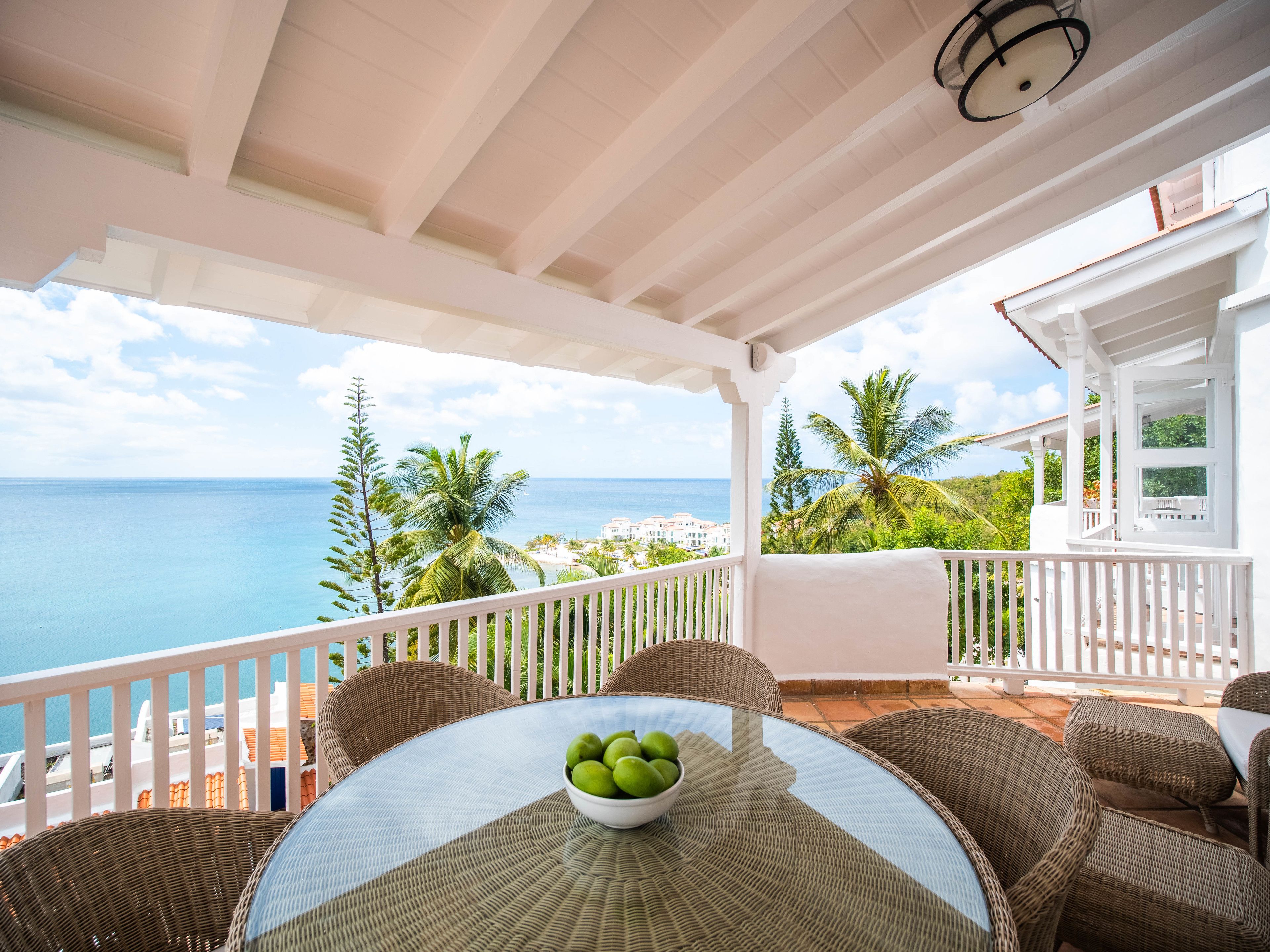 Un espace extérieur aménagé, abrité et doté de mobilier en osier, avec une table sur laquelle se trouve un saladier rempli de fruits, offrant une vue sur la côte, avec des palmiers et des bâtiments.