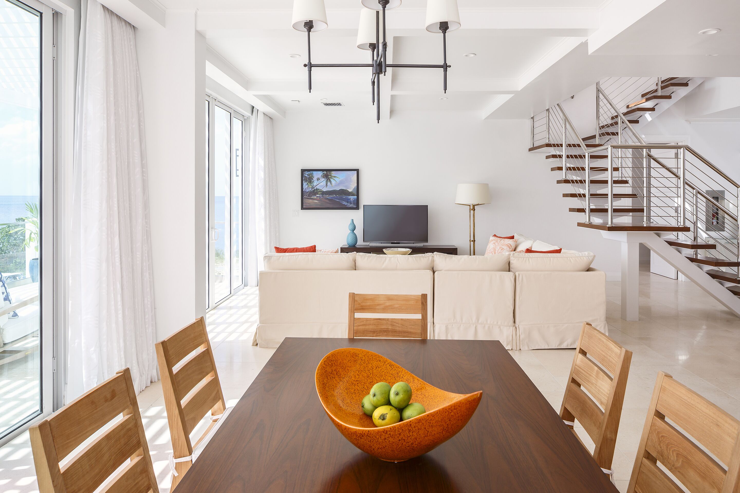 Una sala de estar moderna con una mesa de comedor, sillas de madera, un ventanal grande, un conjunto de sofás, un televisor y una escalera.
