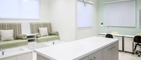 A modern, well-lit room with a kitchenette, a seating area with a sofa and cushions, a desk with a chair, and a large window with blinds.