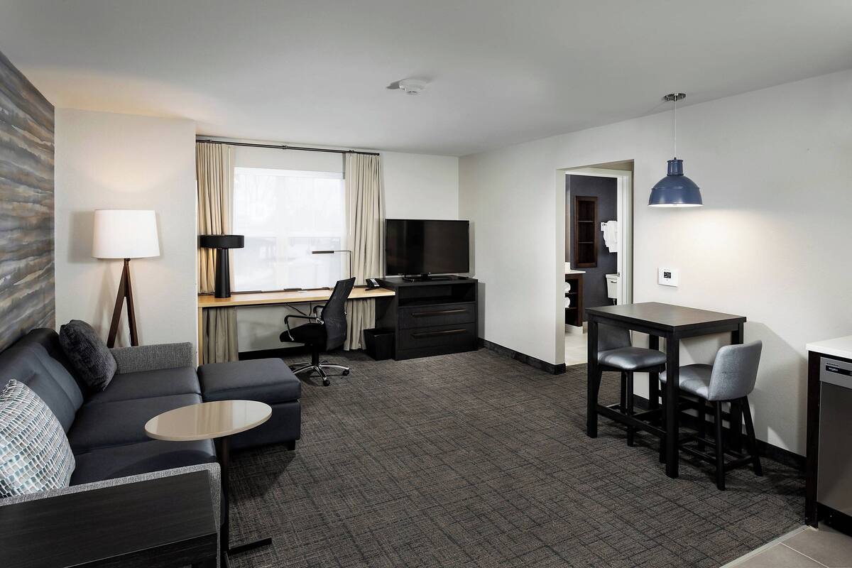 A modern hotel room with a sofa, a small round table, a desk with a chair, a television, and a dining area with a table and chairs.