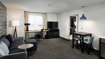 A modern hotel room with a sofa, a small round table, a desk with a chair, a television, and a dining area with a table and chairs.