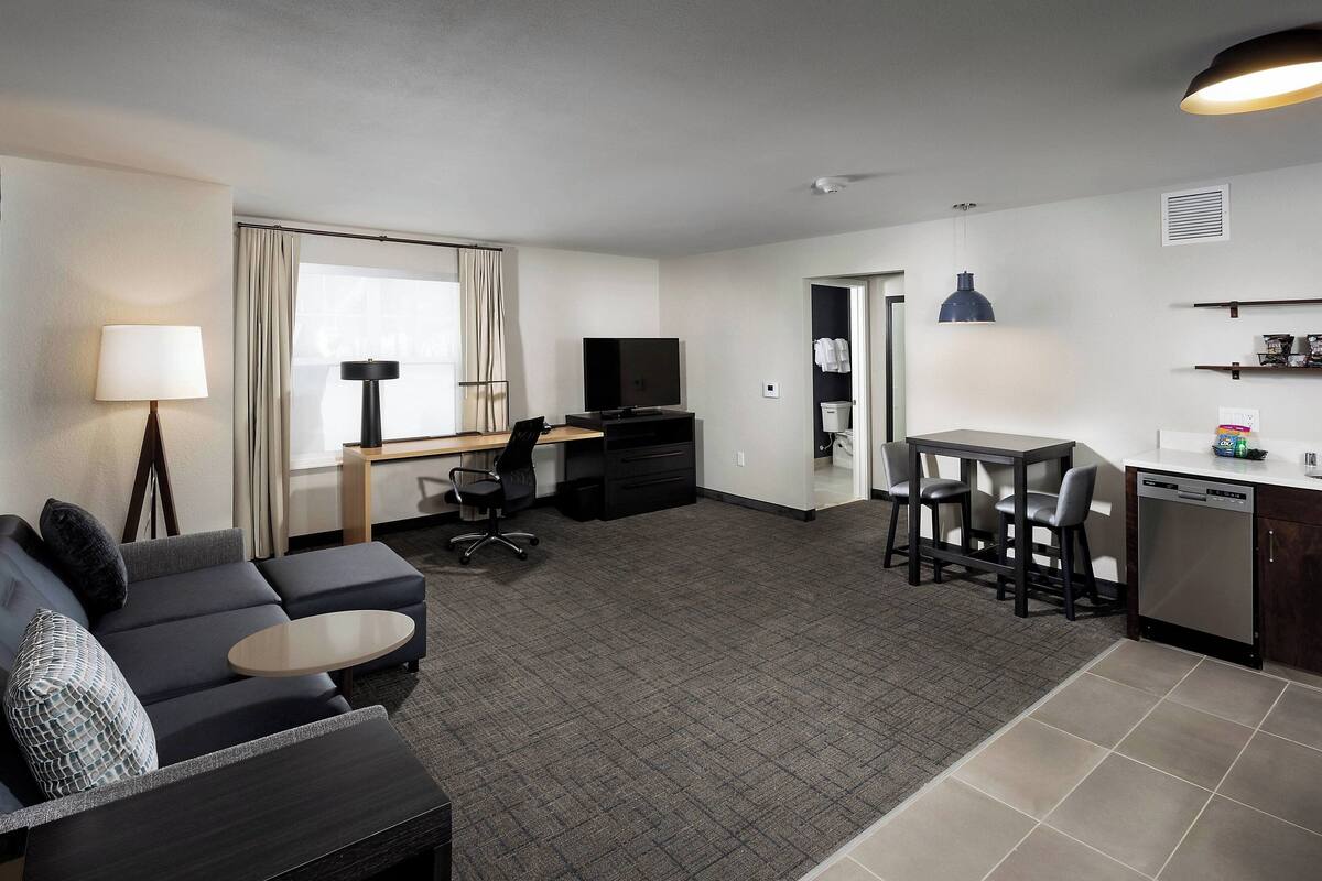 A modern hotel room with a living area, a dining table, a sofa, a TV, and a kitchen area.