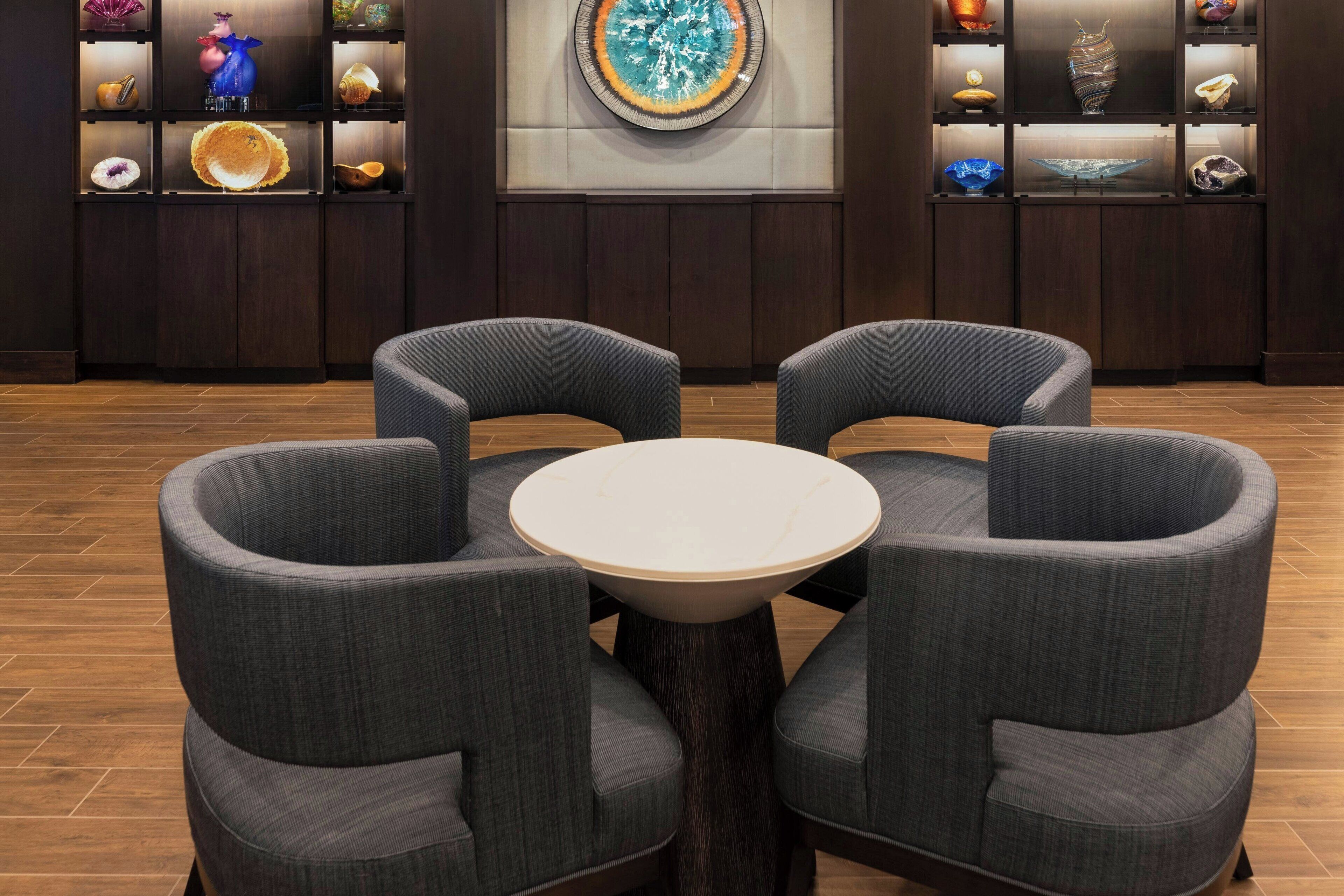 A lobby with a round table surrounded by four grey armchairs, wooden flooring, and a wall with shelves displaying various decorative items.