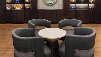 A lobby with a round table surrounded by four grey armchairs, wooden flooring, and a wall with shelves displaying various decorative items.