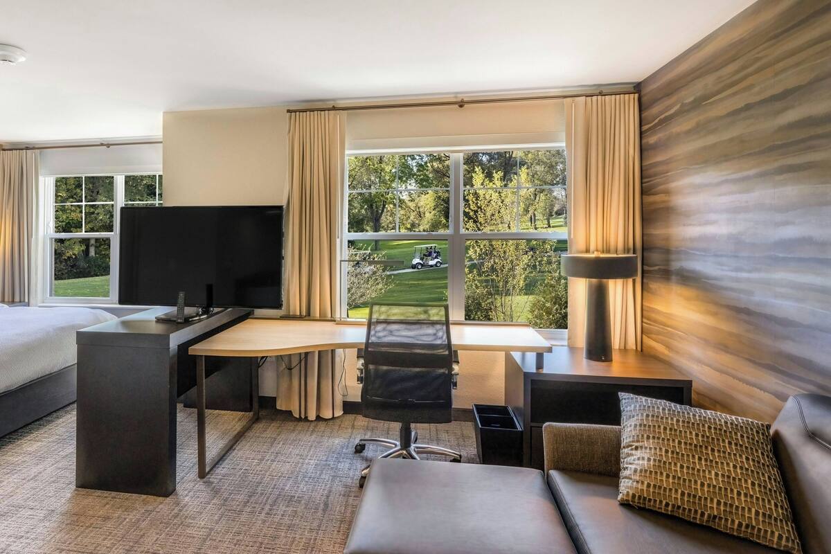 A modern hotel room with a large bed, a desk with a chair, a television, and a view of a golf course.