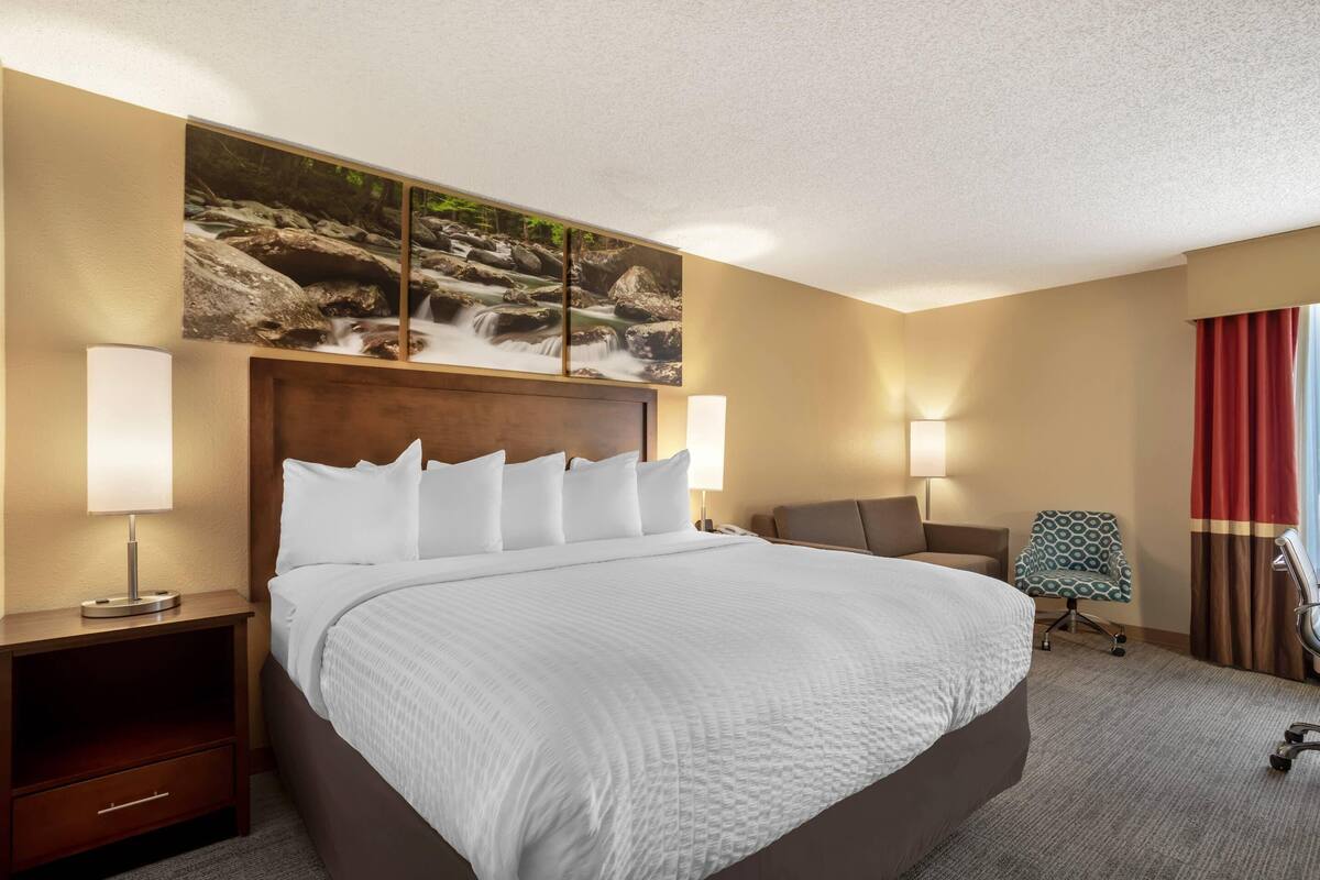 A hotel room with a large bed, two bedside tables with lamps, a chair, a desk, and a painting of a waterfall on the wall.