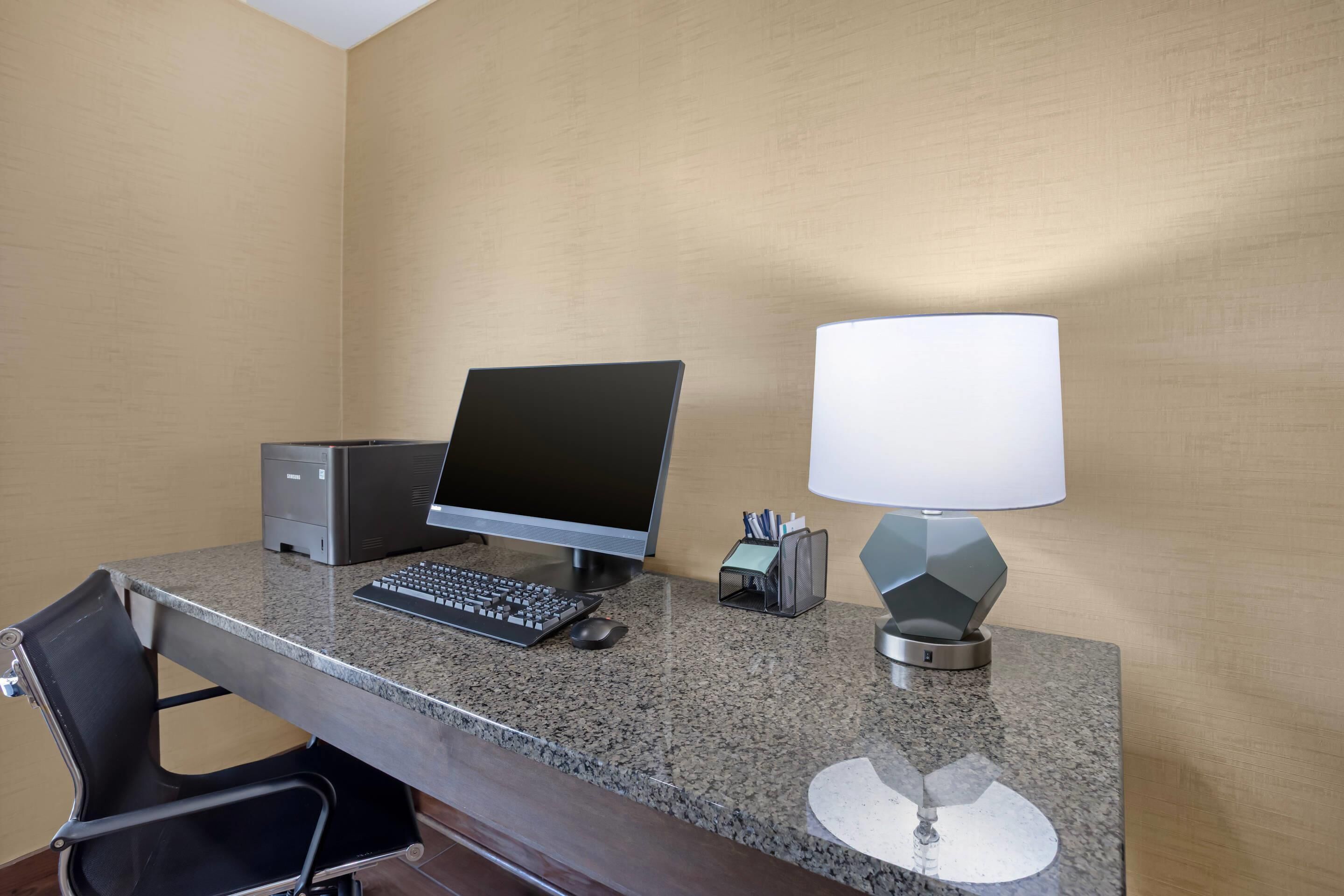 A hotel desk with a desktop computer, keyboard, mouse, lamp, and chair.