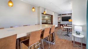 A modern hotel lobby with a long communal table, bar stools, a small bar area, and a television screen. - Clarion Pointe Cherokee (Cherokee)