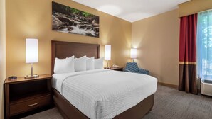 A hotel room with a large bed, two bedside tables with lamps, a nightstand with a drawer, a painting on the wall, and a window with curtains. - Clarion Pointe Cherokee (Cherokee)