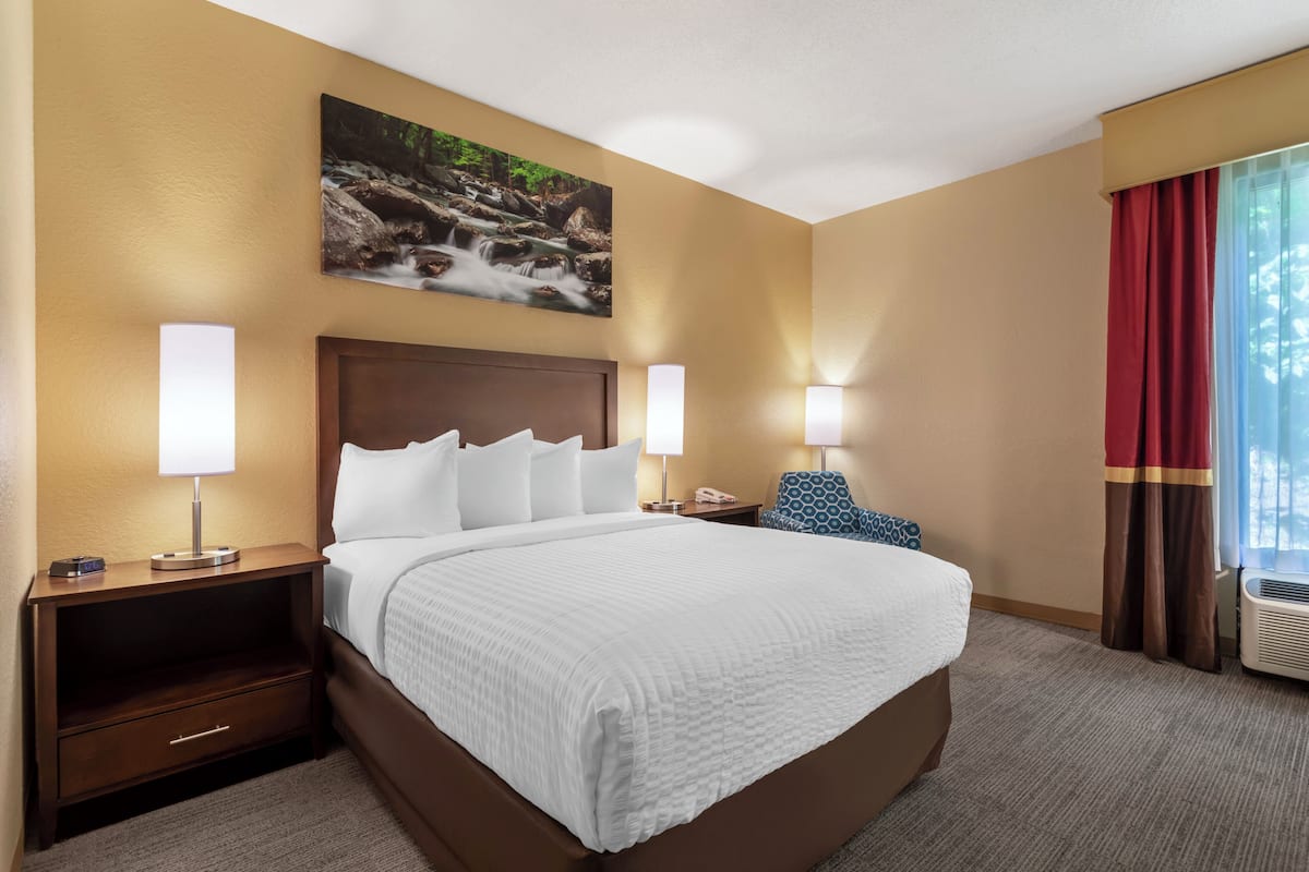 A hotel room with a large bed, two bedside tables with lamps, a nightstand with a drawer, a painting on the wall, and a window with curtains.