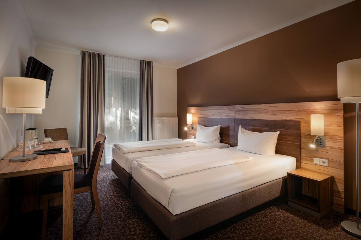 A hotel room with two beds, a desk, a chair, a lamp, and a nightstand.
