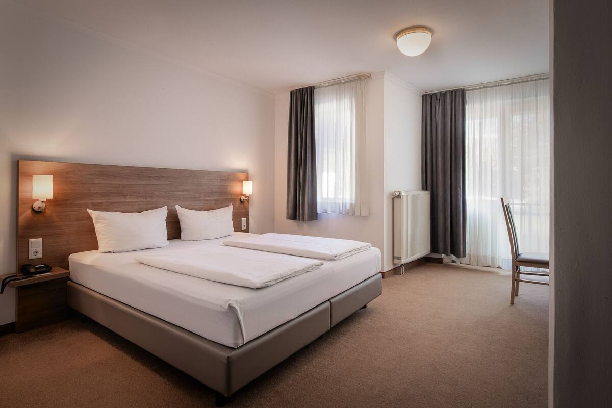 A hotel room with a large bed, bedside lamps, a chair, and a radiator.