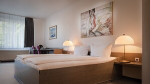 Une chambre d’hôtel avec un grand lit, deux lampes de chevet, un bureau avec une chaise, un radiateur et une fenêtre avec des rideaux.