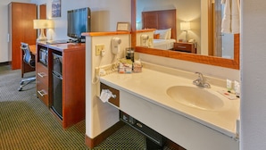A hotel room with a desk, a television, a minibar, a large mirror, a sink, and a towel rack.