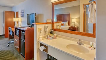 Ein Hotelzimmer mit Schreibtisch, Fernseher, Minibar, großem Spiegel, Waschbecken und Handtuchhalter.