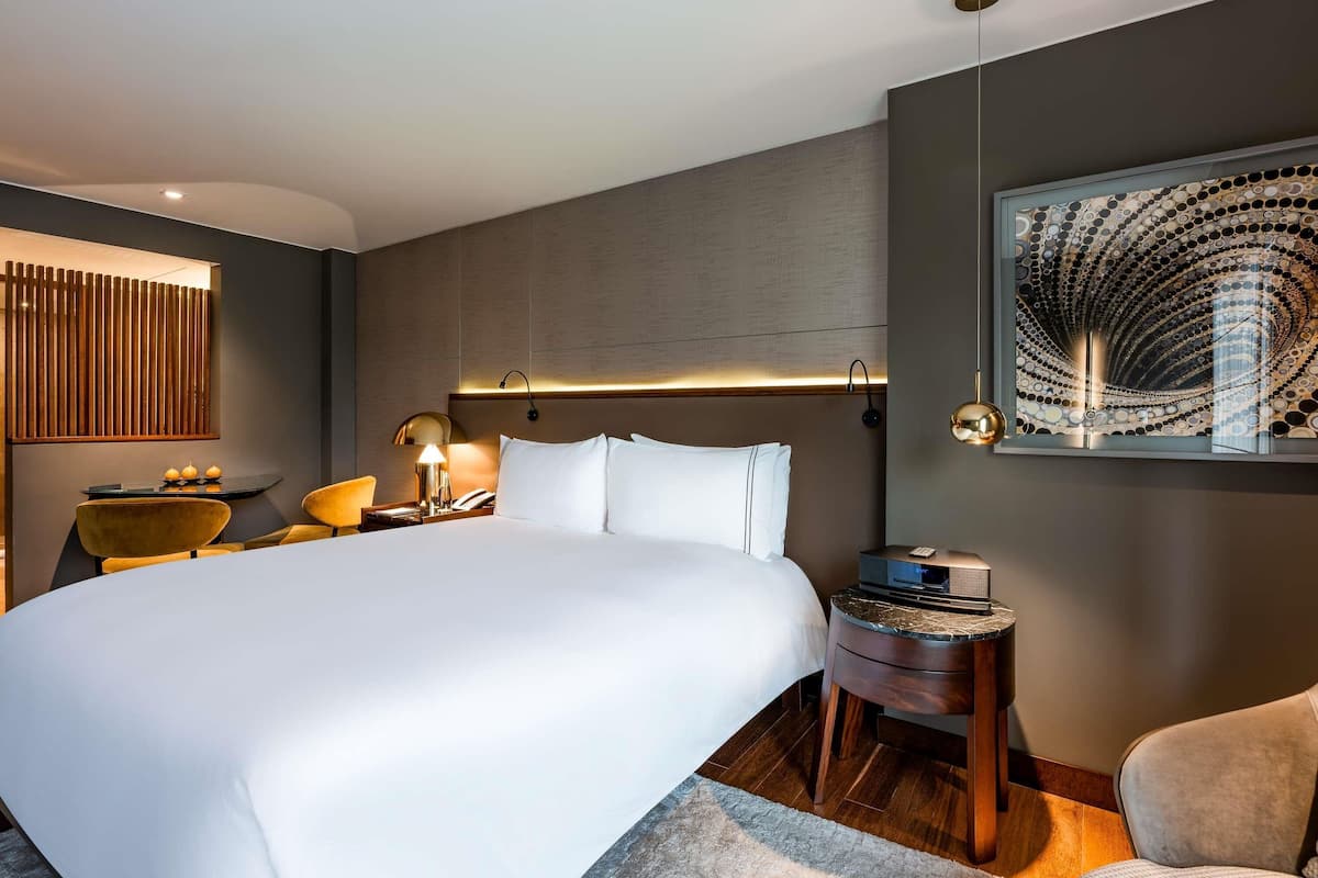 A modern hotel room with a large bed, a bedside table with a lamp, a small side table, and a wall-mounted artwork.