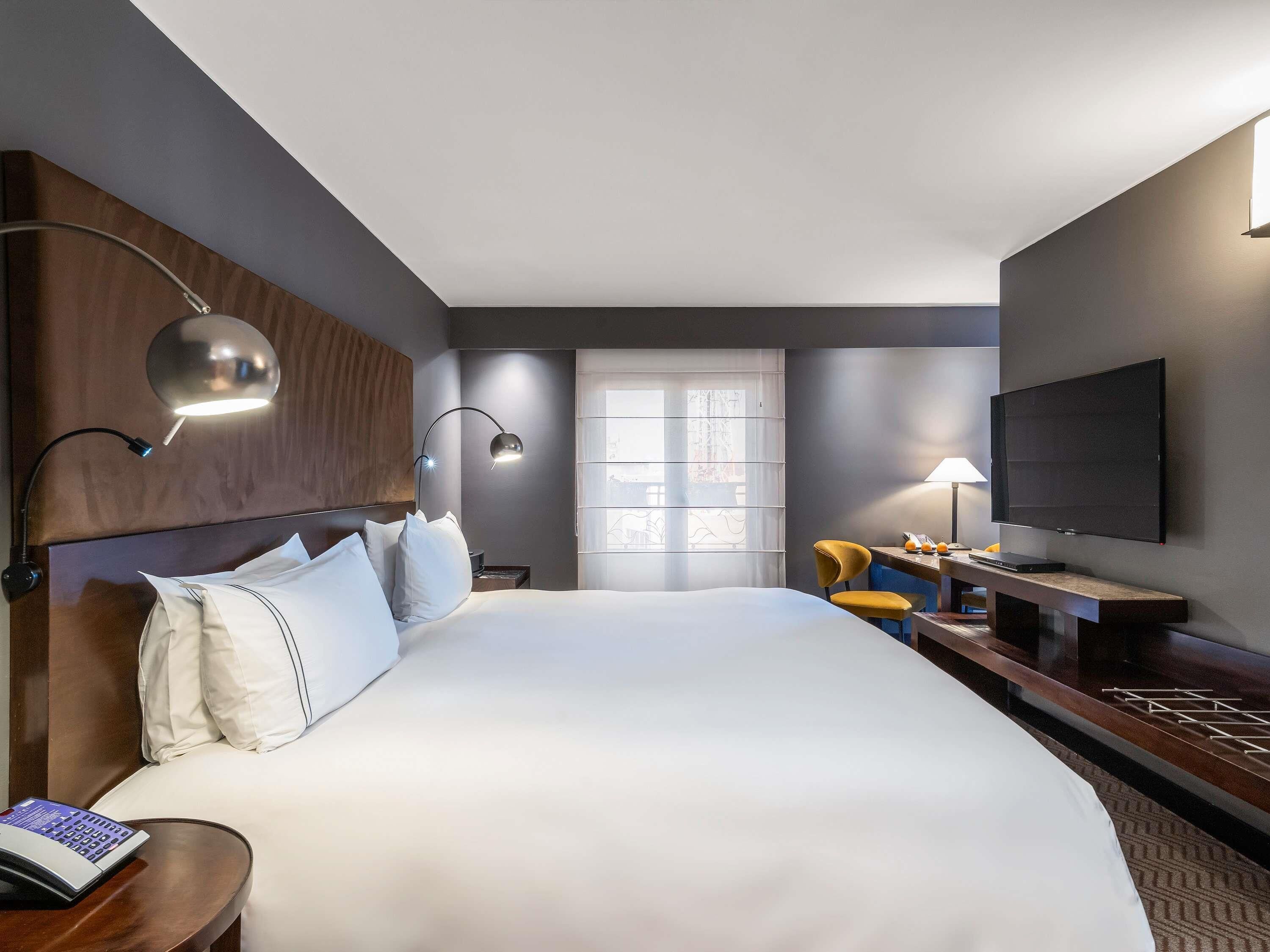 A hotel room with a large bed, bedside lamps, a flat-screen TV, a desk with a lamp, and a window with blinds.