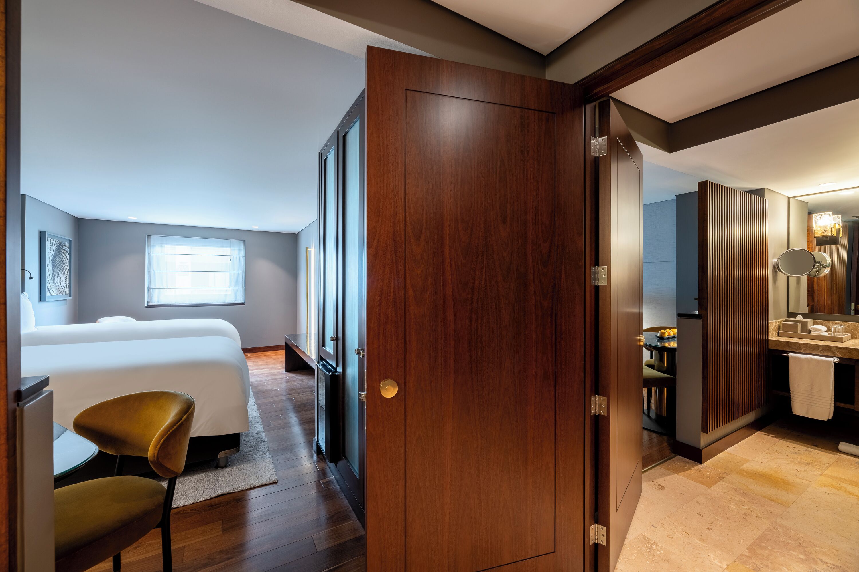 A bedroom with a large bed, a chair, and a wooden door leading to a bathroom with a sink and mirror.
