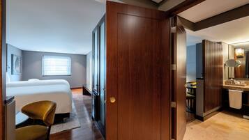 A bedroom with a large bed, a chair, and a wooden door leading to a bathroom with a sink and mirror.