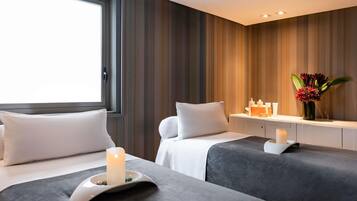 Ein Hotelzimmer mit zwei Betten, einem Nachttisch mit Pflegeprodukten, einer Vase mit Blumen und einem Fenster mit Vorhängen.