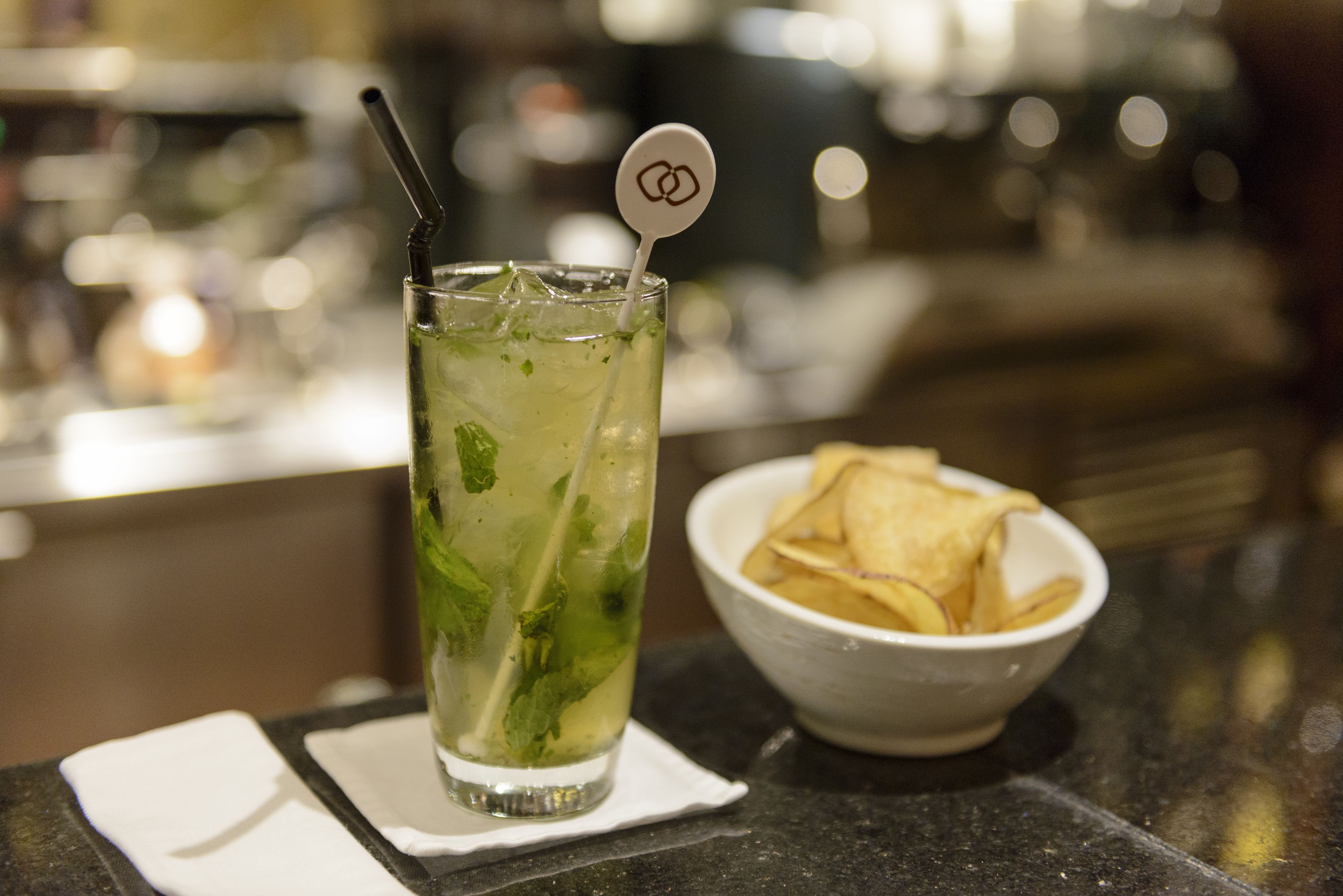 Un vaso de bebida helada con pajita y una ramita de menta, un cuenco de patatas fritas y una servilleta sobre una barra.