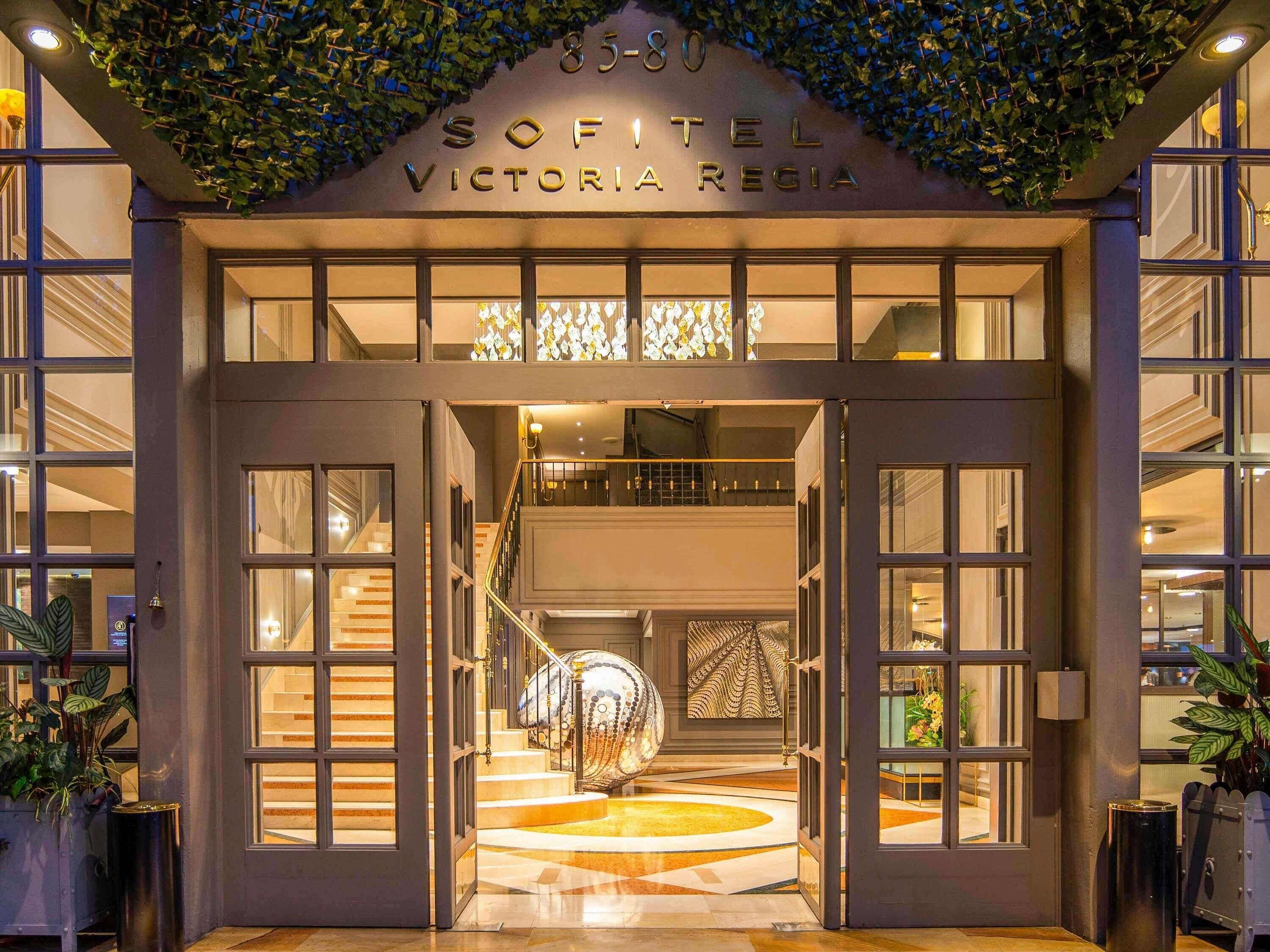 The entrance of Sofitel Victoria Regina with a grand staircase, large windows, and a decorative ceiling.
