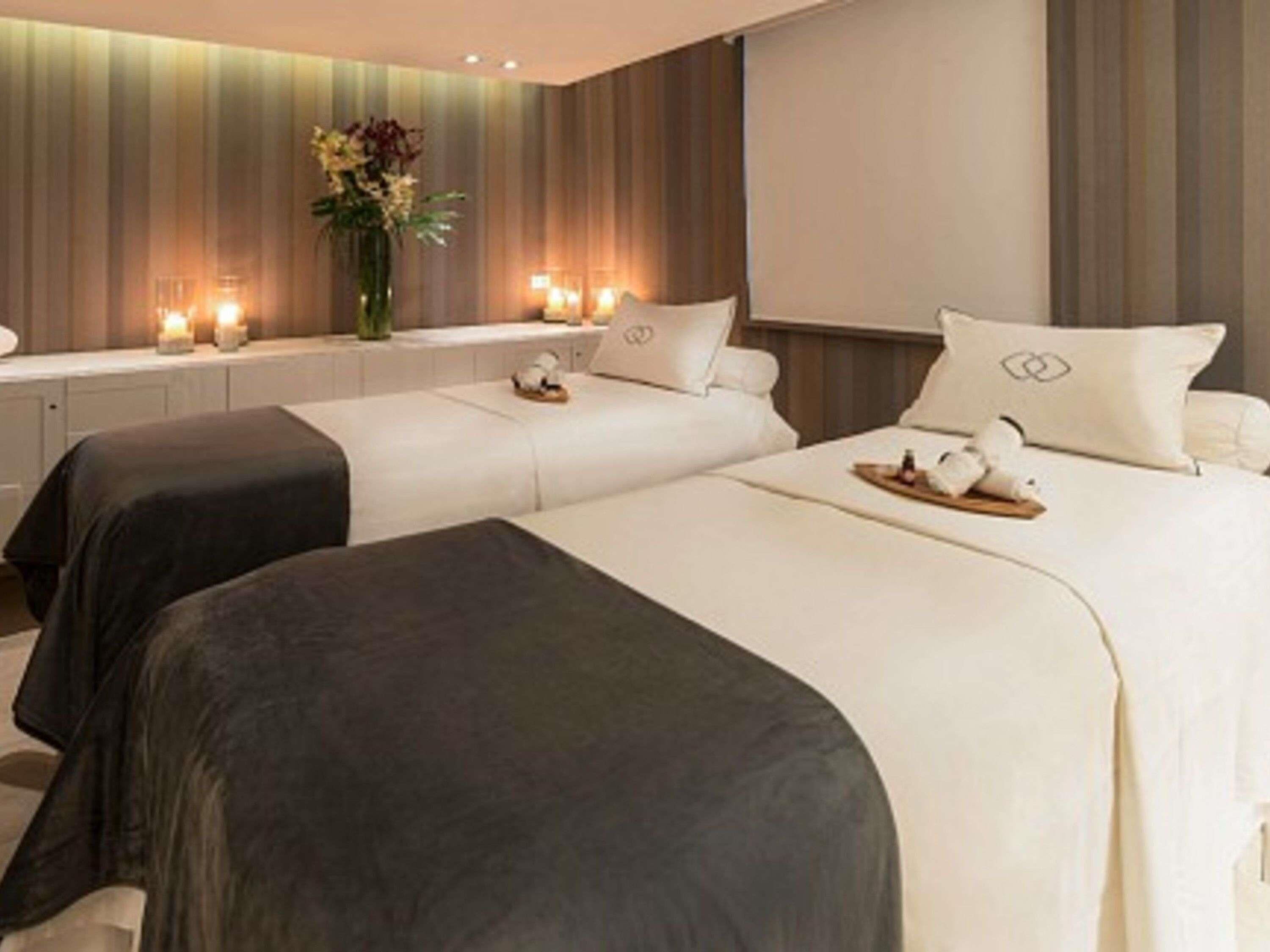 A hotel room with two beds, a bedside table, a vase with flowers, and a wall-mounted TV.