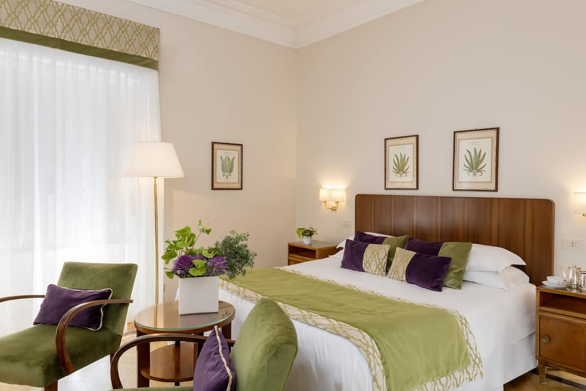 A hotel room with a bed, two chairs, a small table, a lamp, and framed botanical prints on the wall.