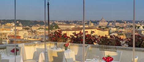 A rooftop restaurant with white wicker furniture, tables set with white tablecloths, and red flowers. A view of a cityscape with historic buildings is visible in the background.