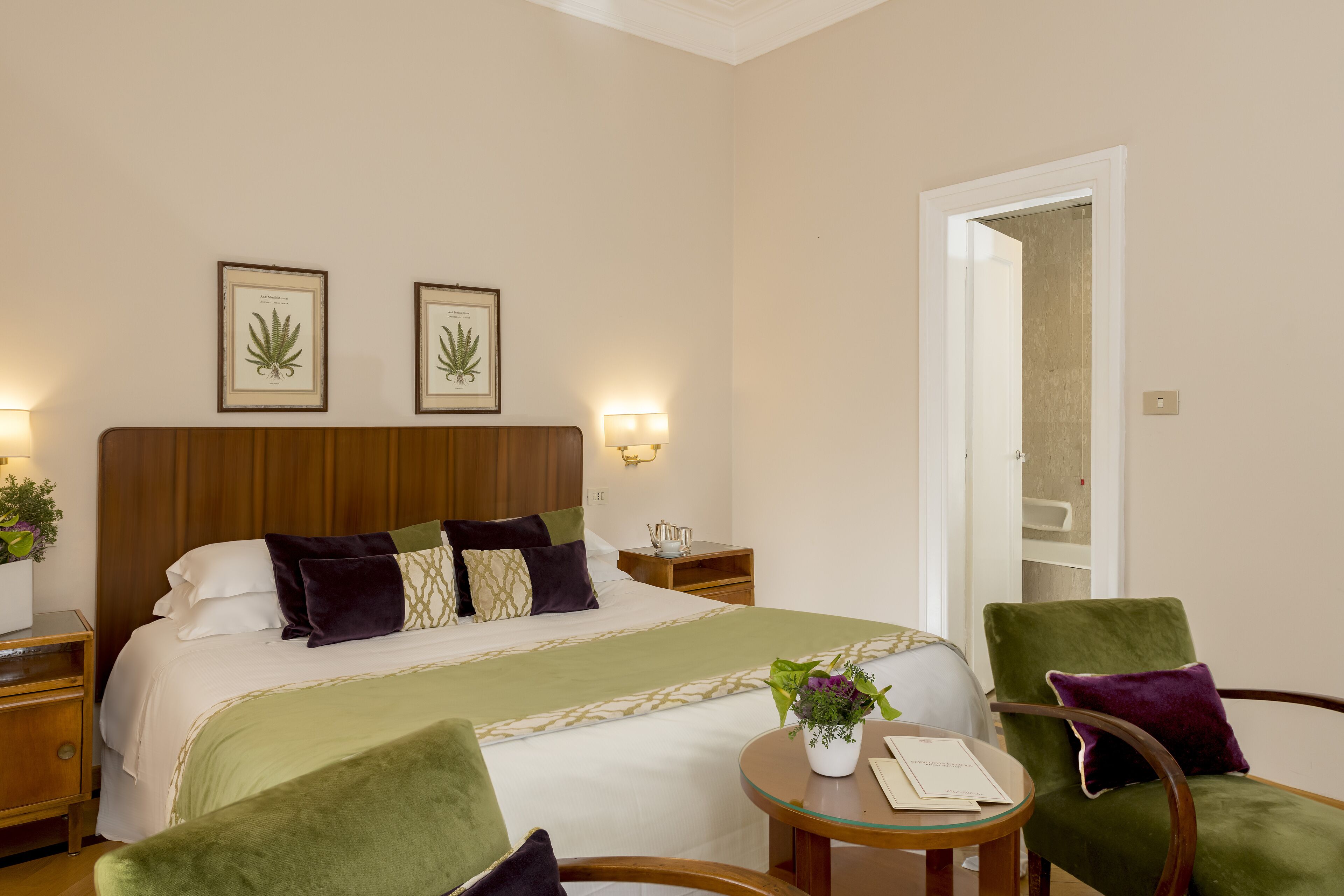 A hotel room with a large bed, two framed botanical prints, a bedside table with a lamp, a small round table with a plant, and two green armchairs.