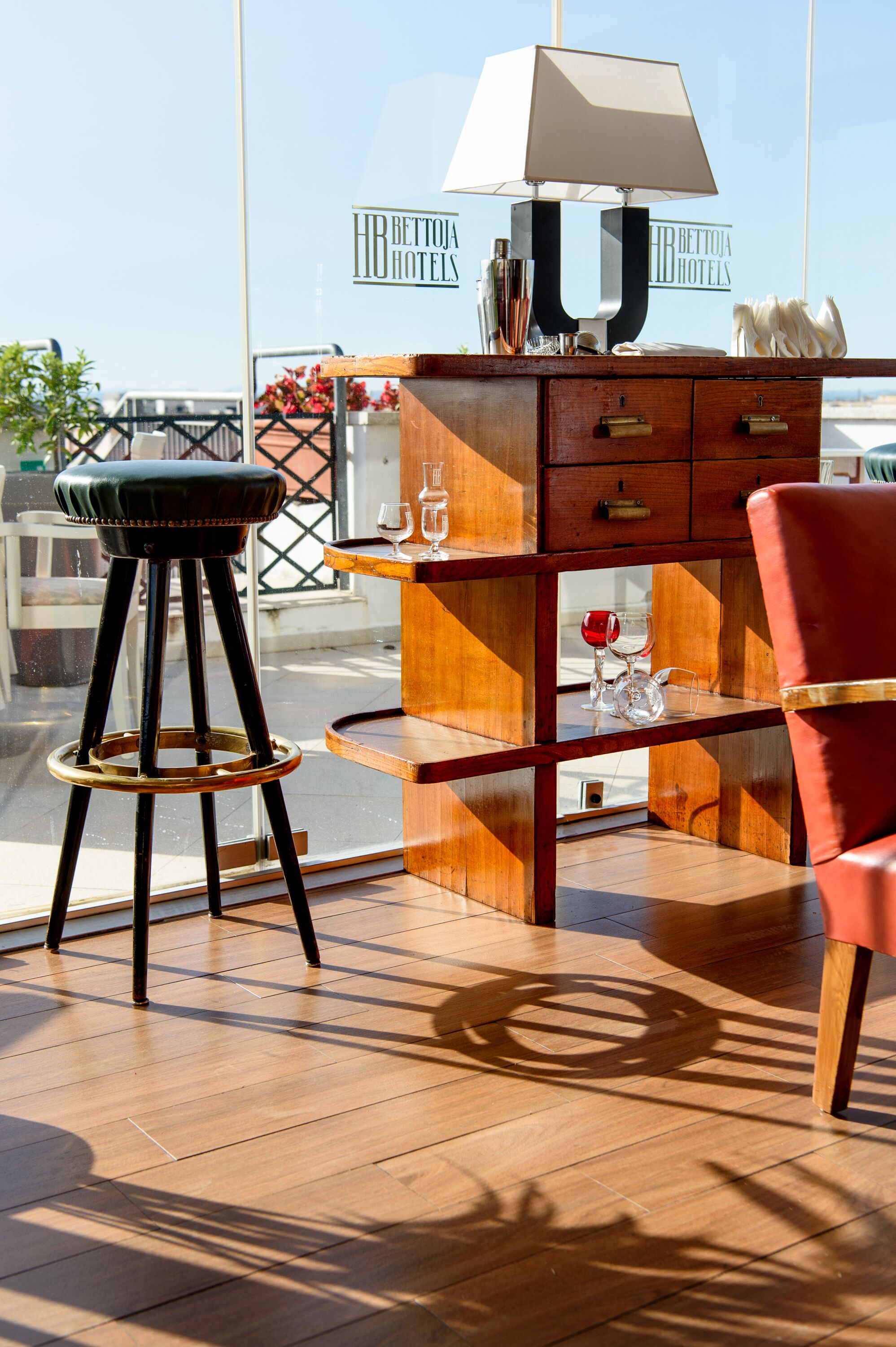 A wooden desk with a lamp, a red chair, and a bar stool in a room with a large window.