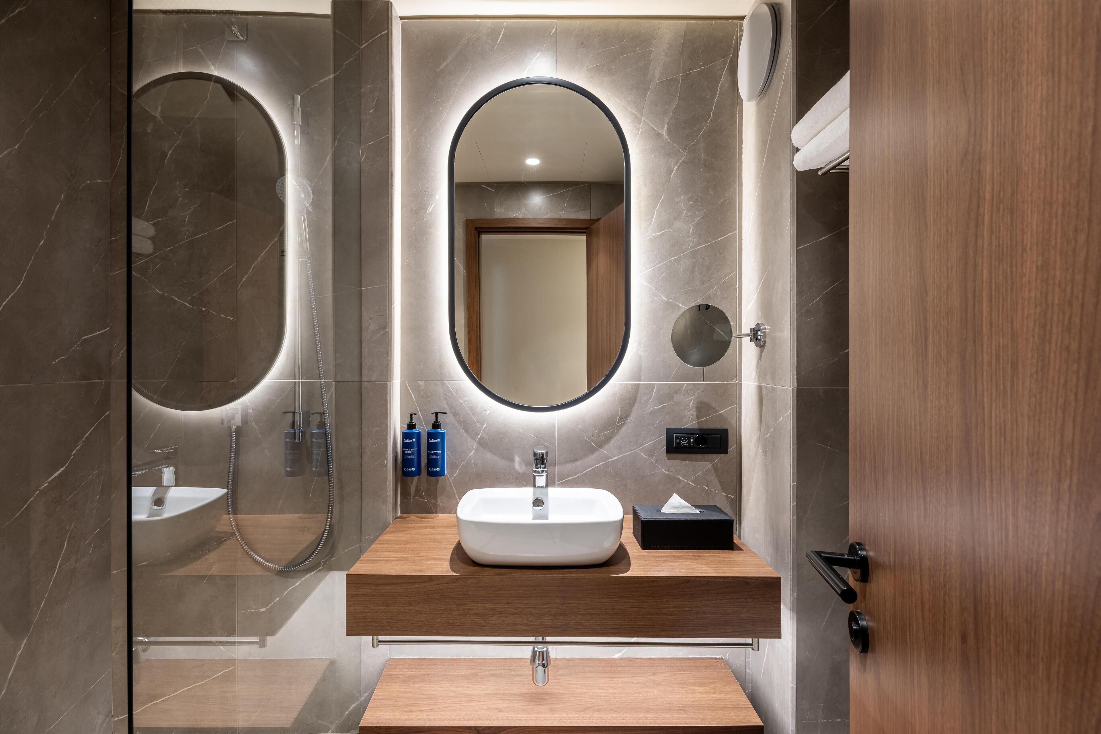 Une salle de bain moderne avec une douche à parois de verre, un miroir rond, un meuble-lavabo en bois et un lavabo intégré.