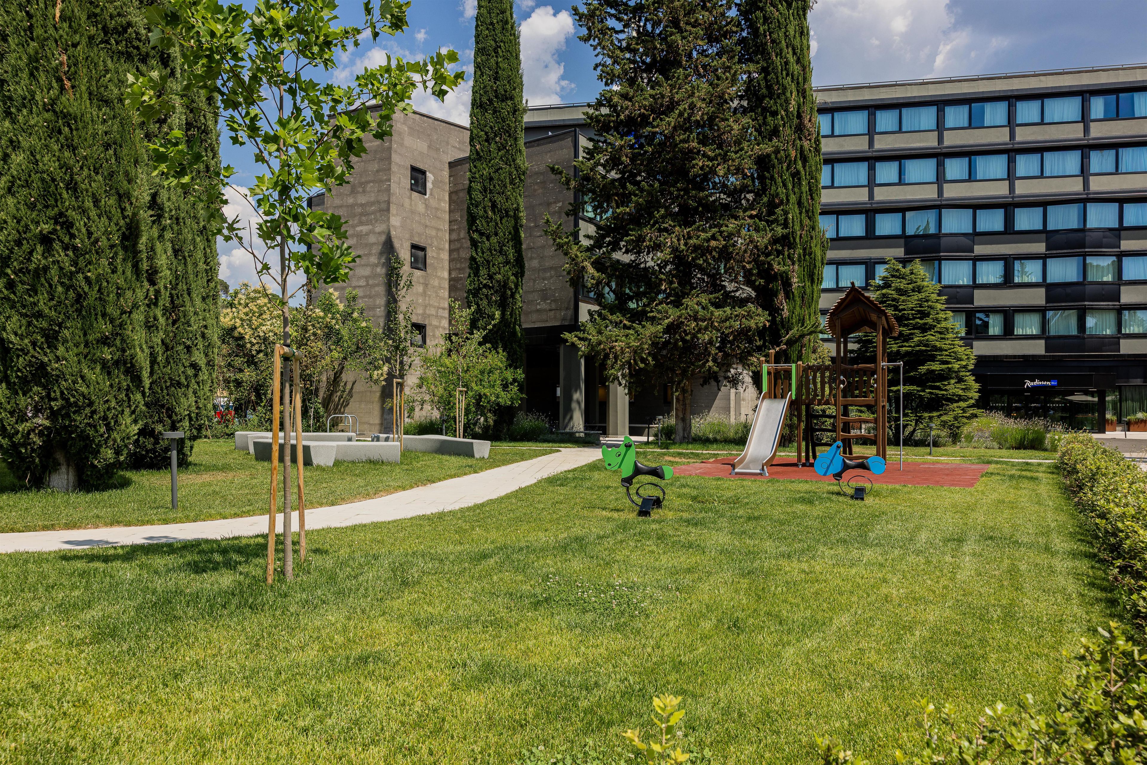 Un parque infantil con columpios y un tobogán, rodeado de césped y árboles, con un edificio al fondo.