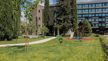 Ein Spielplatz mit Schaukeln und Rutsche, umgeben von Gras und Bäumen, mit einem Gebäude im Hintergrund.
