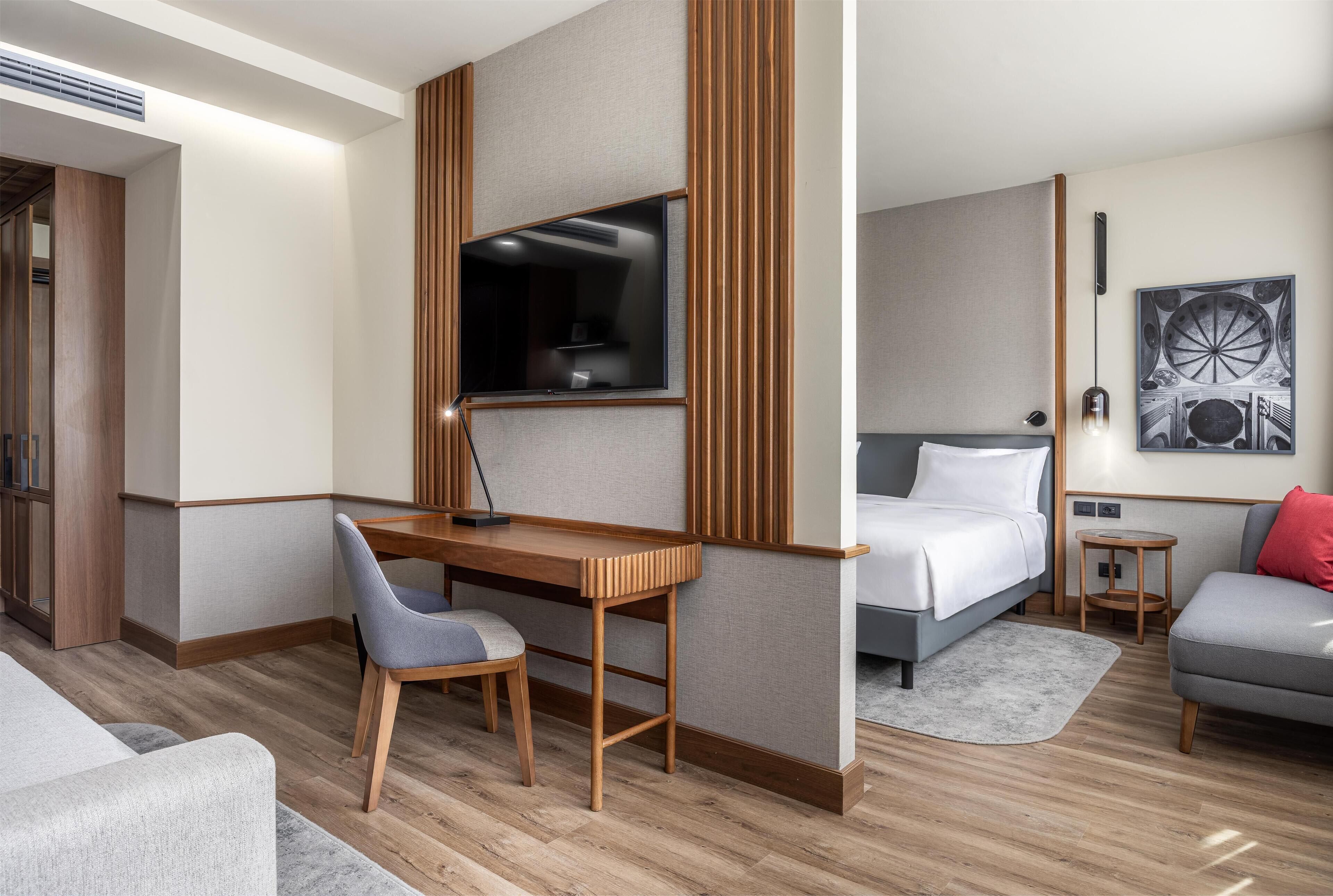 A modern hotel room with a wooden desk, a bed, a grey sofa, and a small round table.
