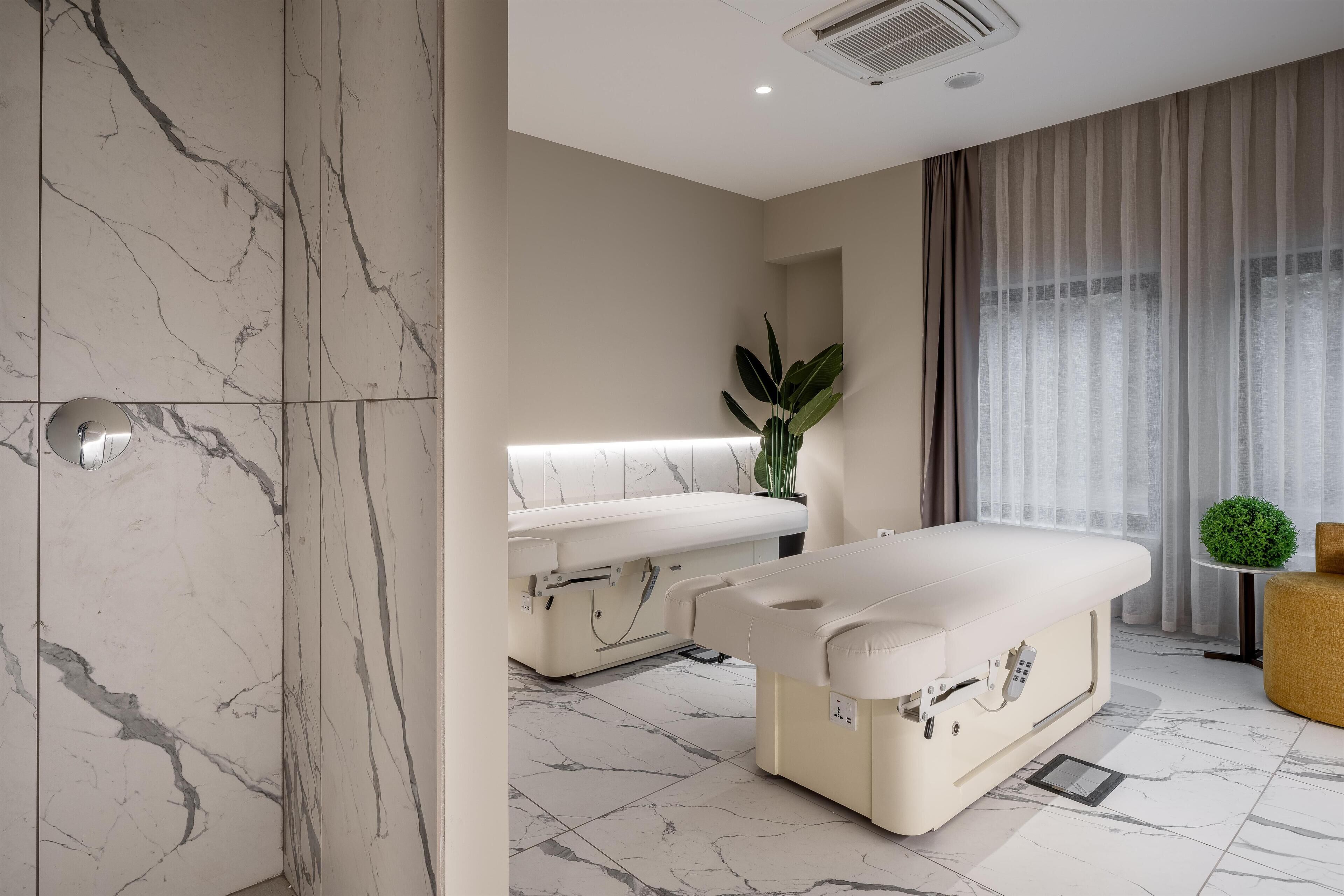 A modern spa room with marble flooring, a massage table, a bench, and a potted plant.