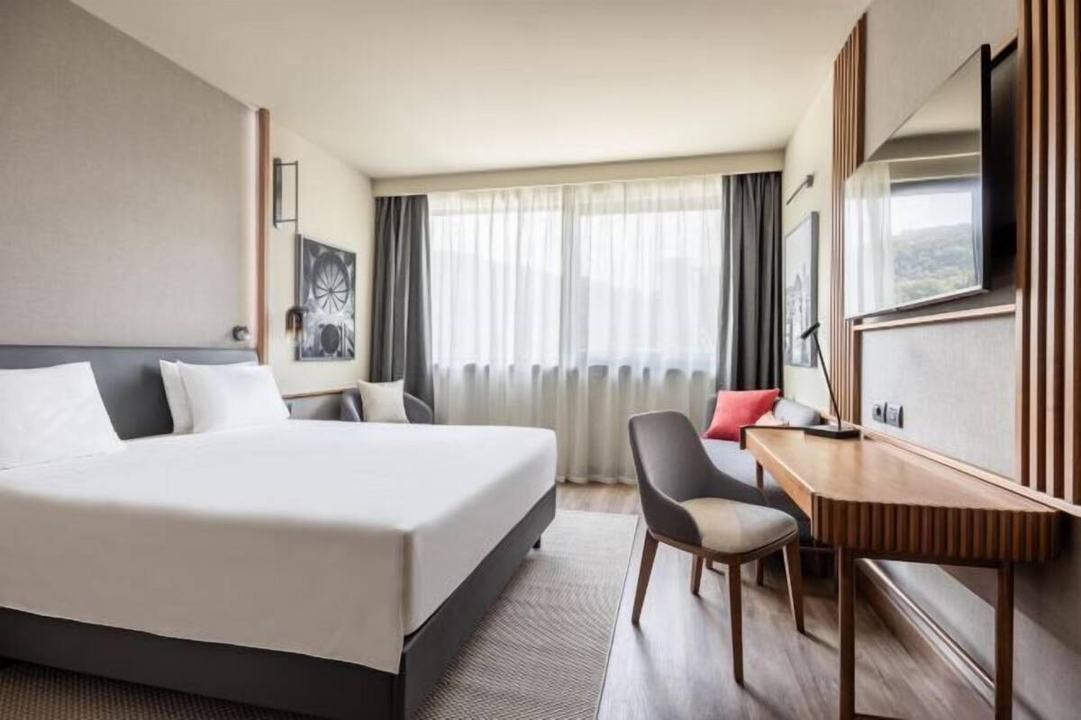 Une chambre d’hôtel avec un grand lit, un bureau, une chaise, une télévision et une fenêtre avec des rideaux.
