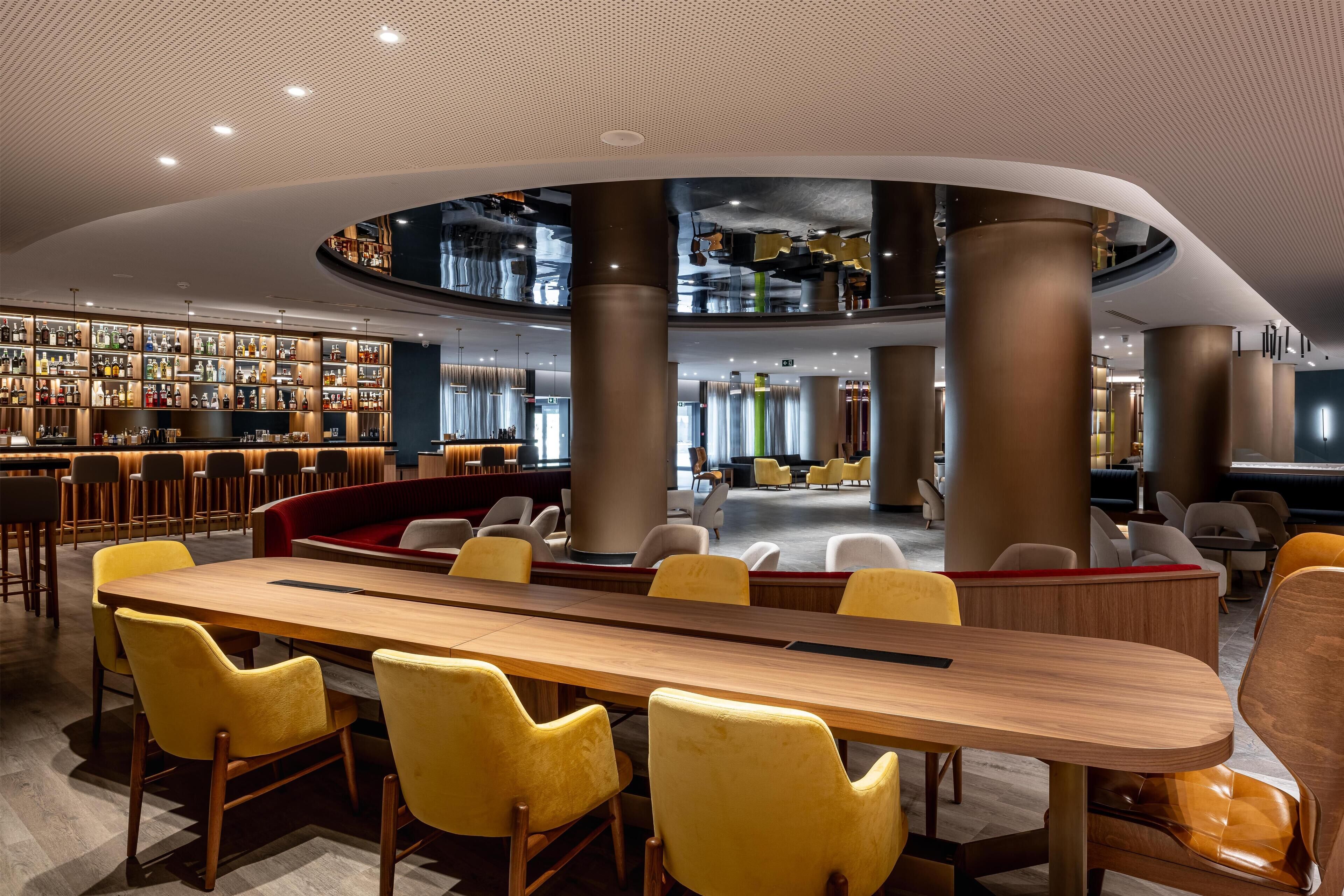A modern lounge area with a long wooden table, yellow chairs, and a bar with shelves of bottles.