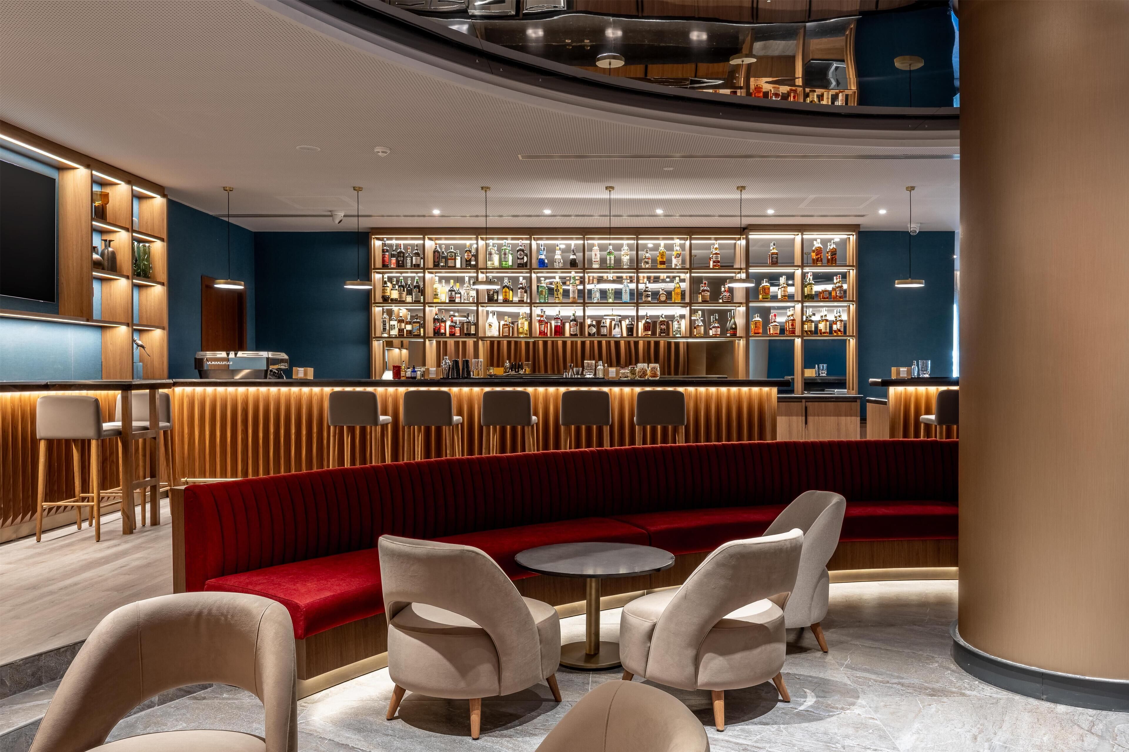 A modern bar area with a red velvet booth, round tables, and a variety of bottles on shelves.