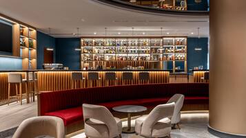 A modern bar area with a red velvet booth, round tables, and a variety of bottles on shelves.