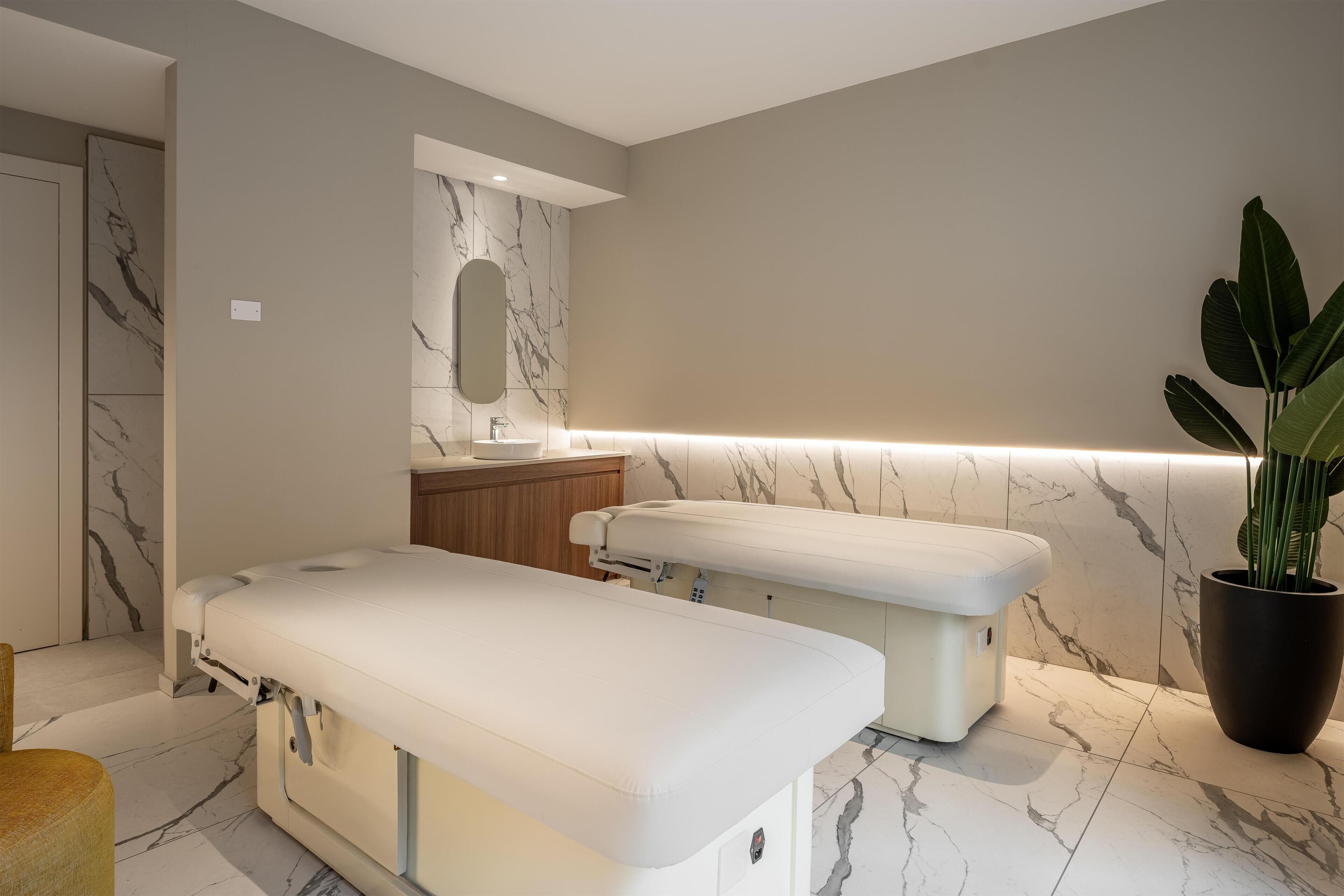 Een moderne spa-ruimte met twee massagetafels, een houten kast, een marmeren muur en een potplant.