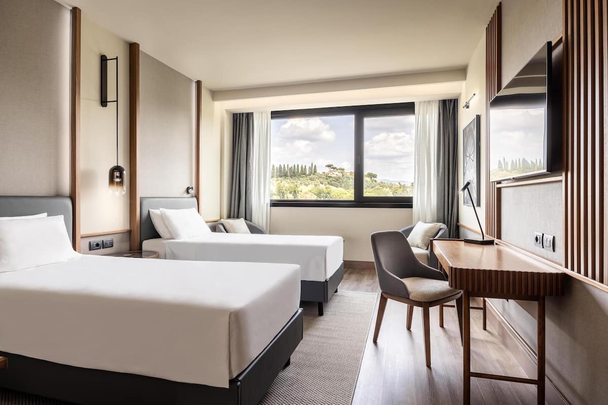 Une chambre d’hôtel avec deux lits, un bureau et une chaise. Une grande fenêtre offre une vue sur les arbres et les collines.