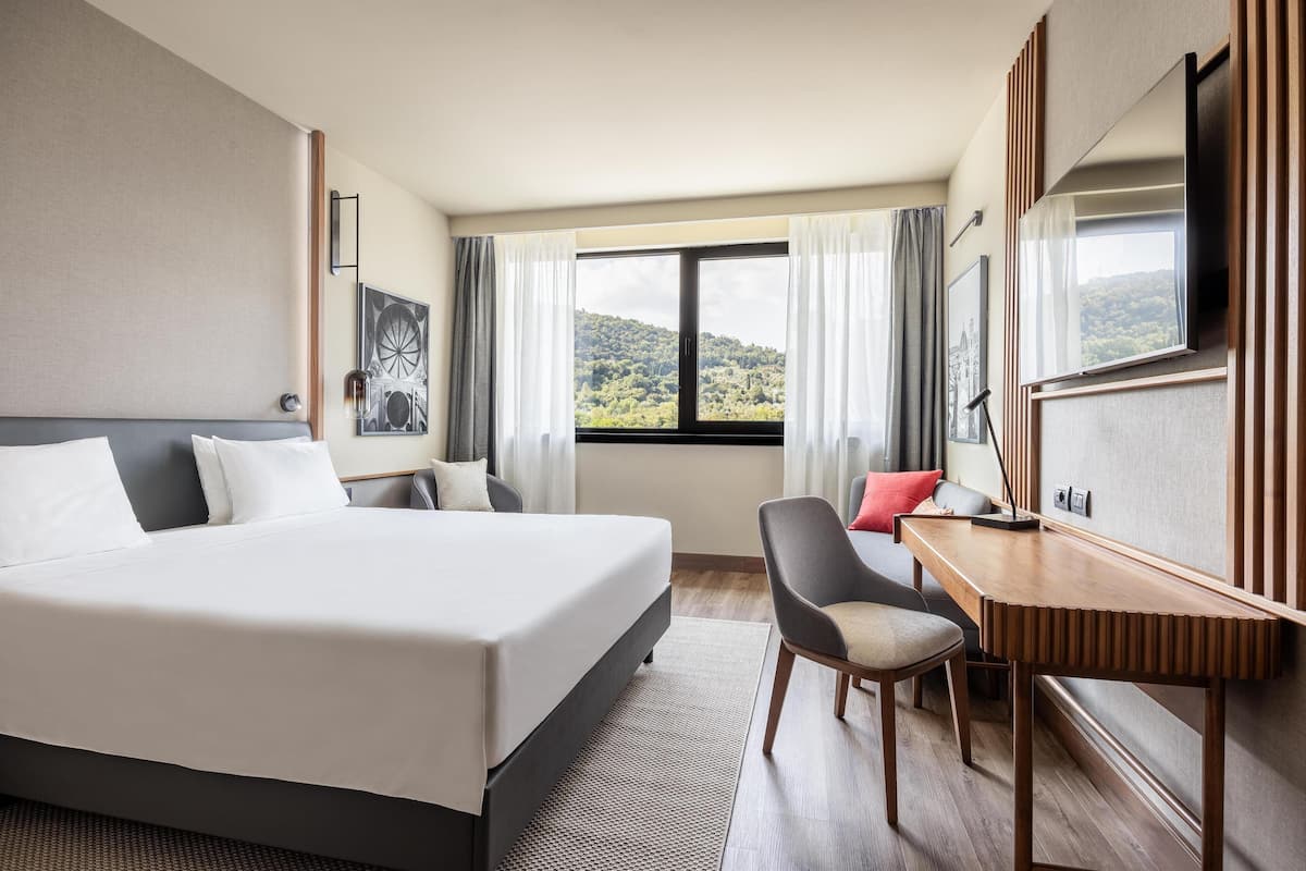 Une chambre d’hôtel avec un grand lit, un bureau, une chaise et une vue sur un paysage montagneux par la fenêtre.