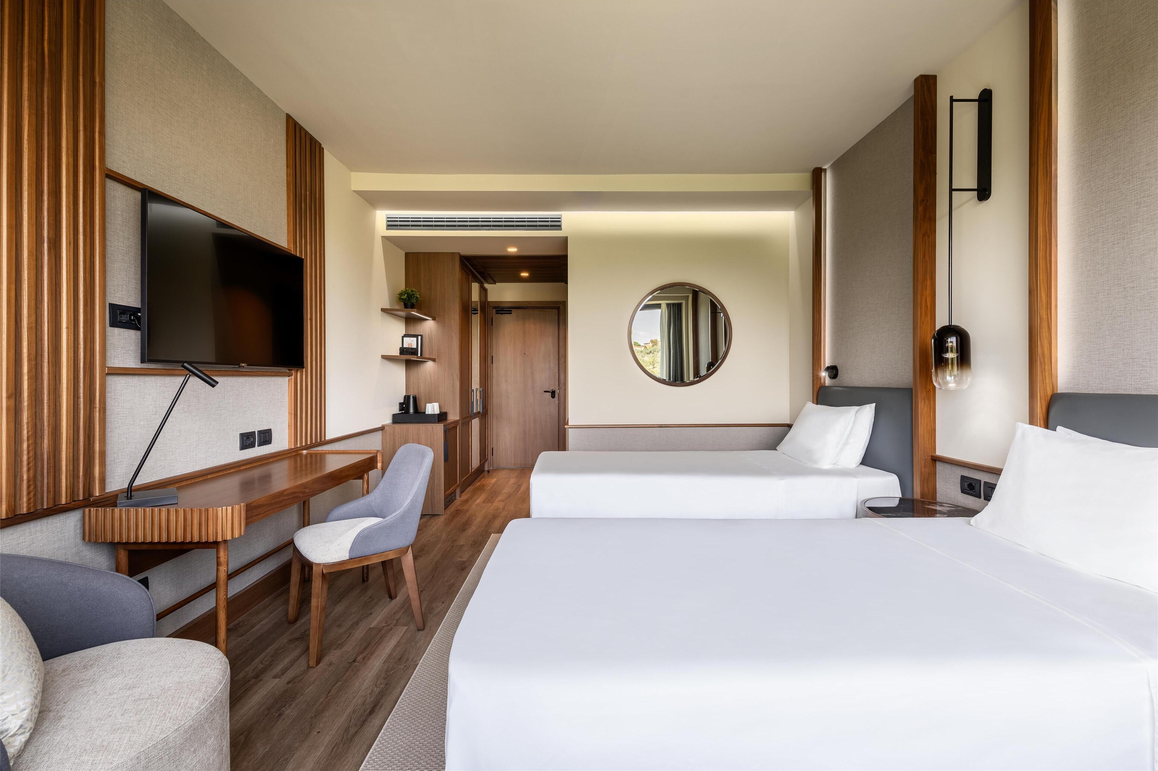 Habitación de hotel con cama, escritorio, silla, televisor y espejo redondo.