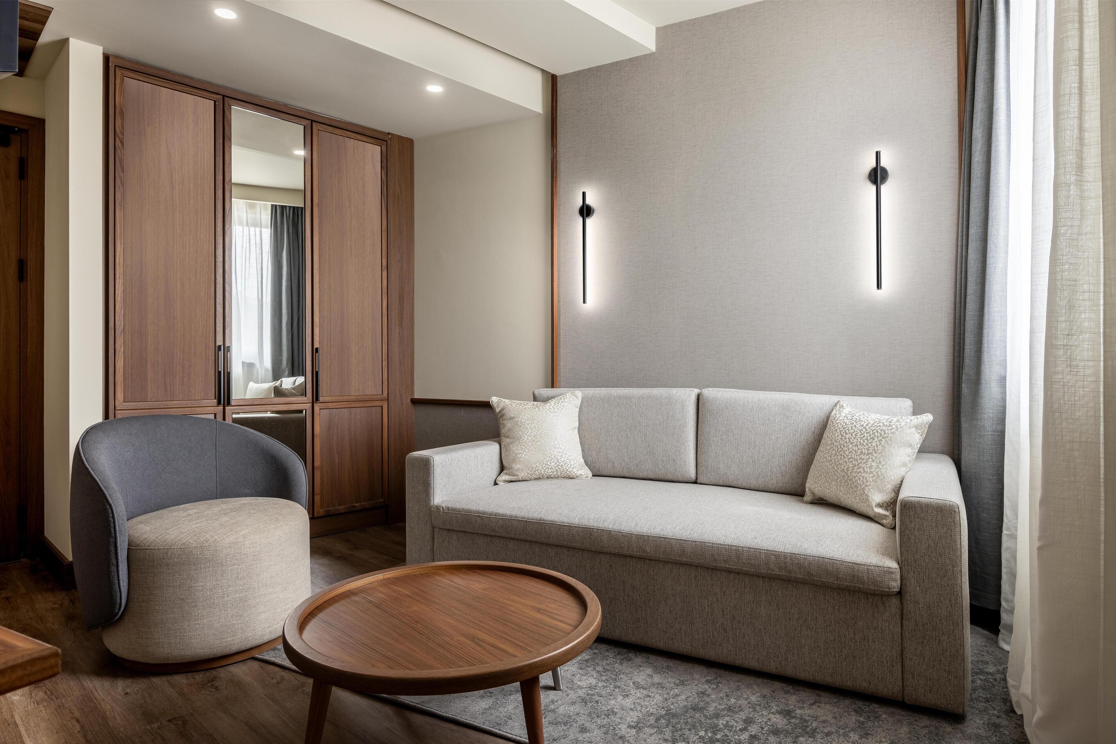 A modern living room with a sofa, a chair, a round wooden table, and a wardrobe with a mirror.