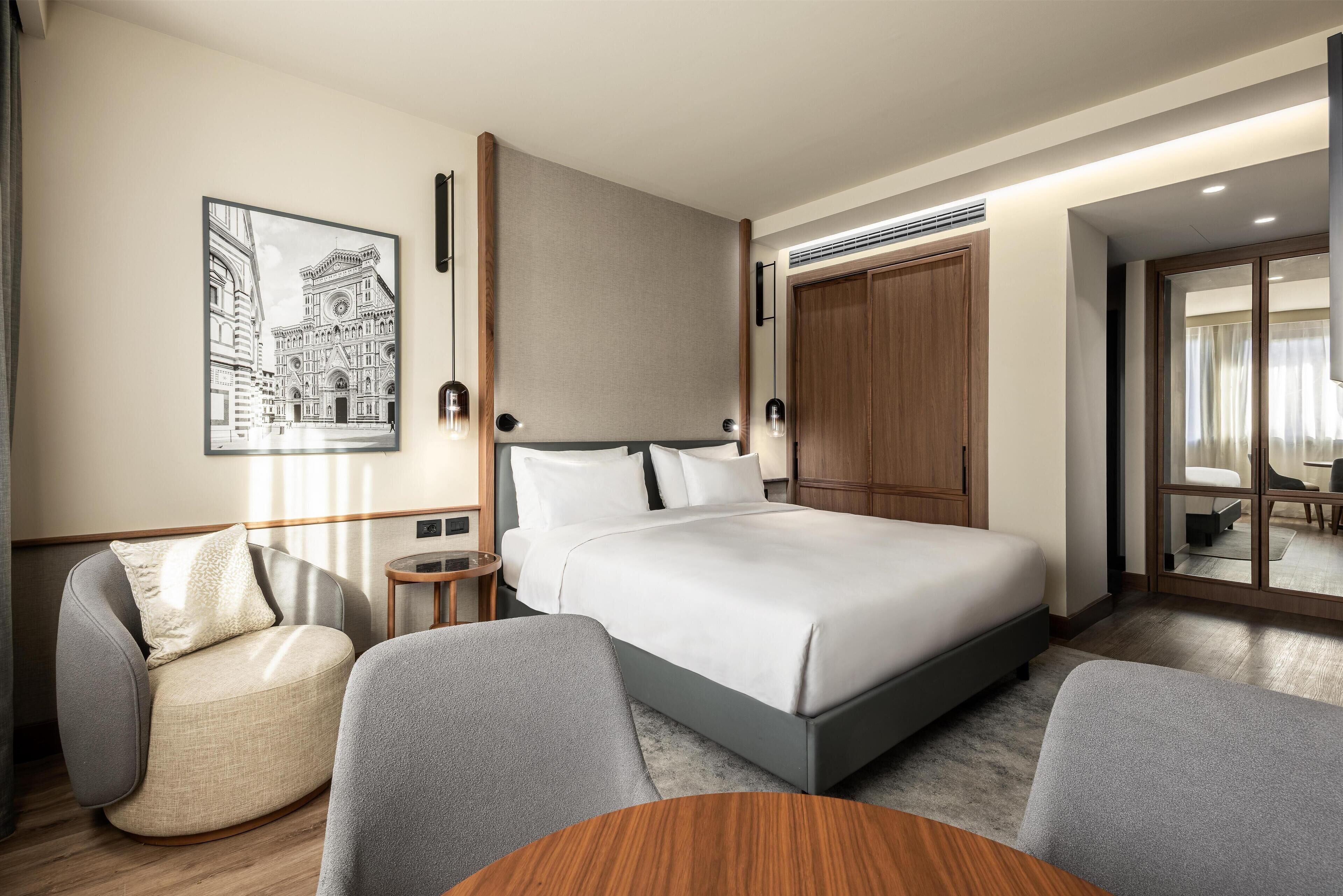 A modern hotel room with a large bed, a seating area with a round table, and a framed artwork on the wall.