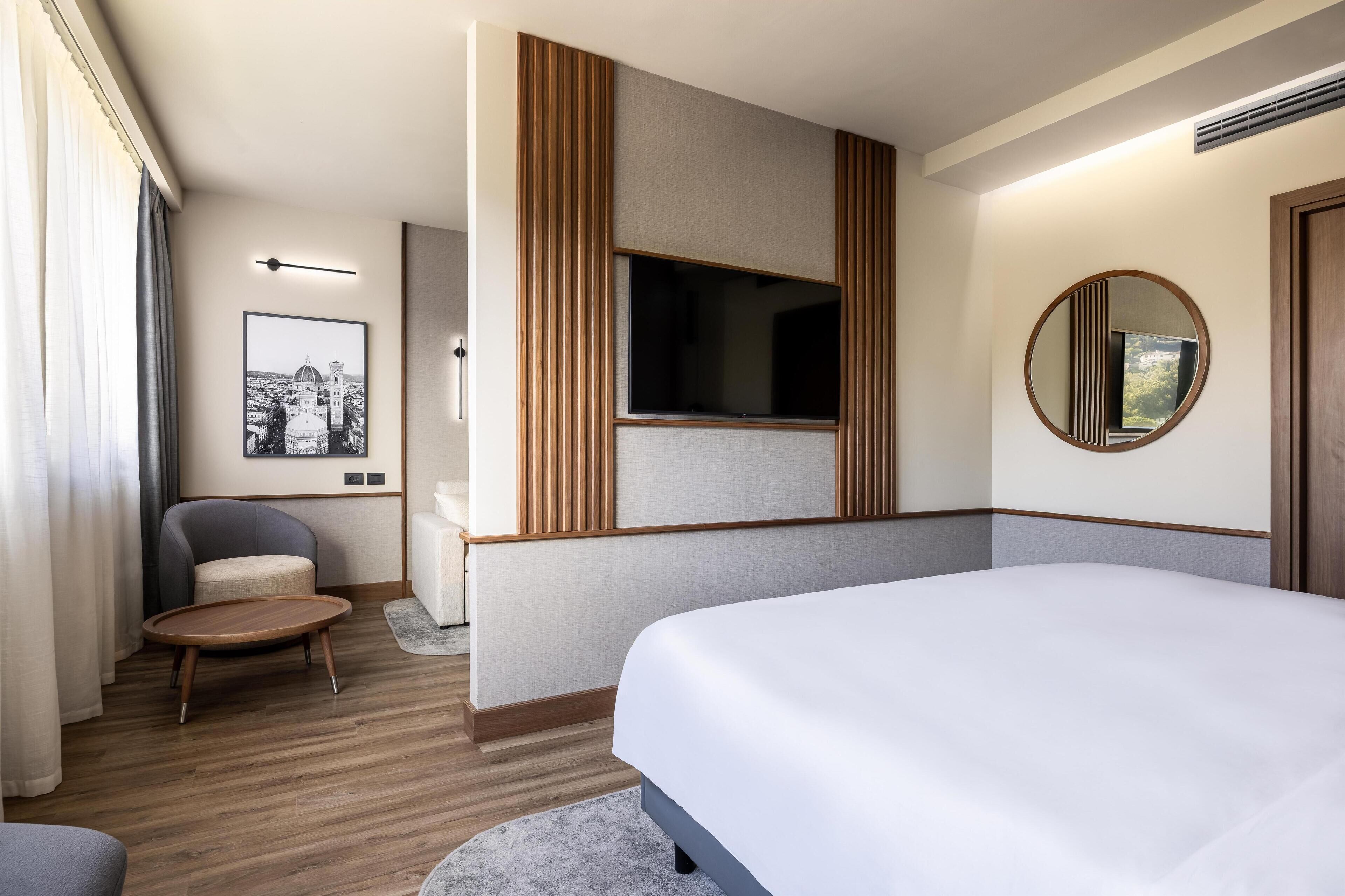 A modern hotel room with a large bed, a flat-screen TV, a round mirror, and a framed cityscape picture on the wall.