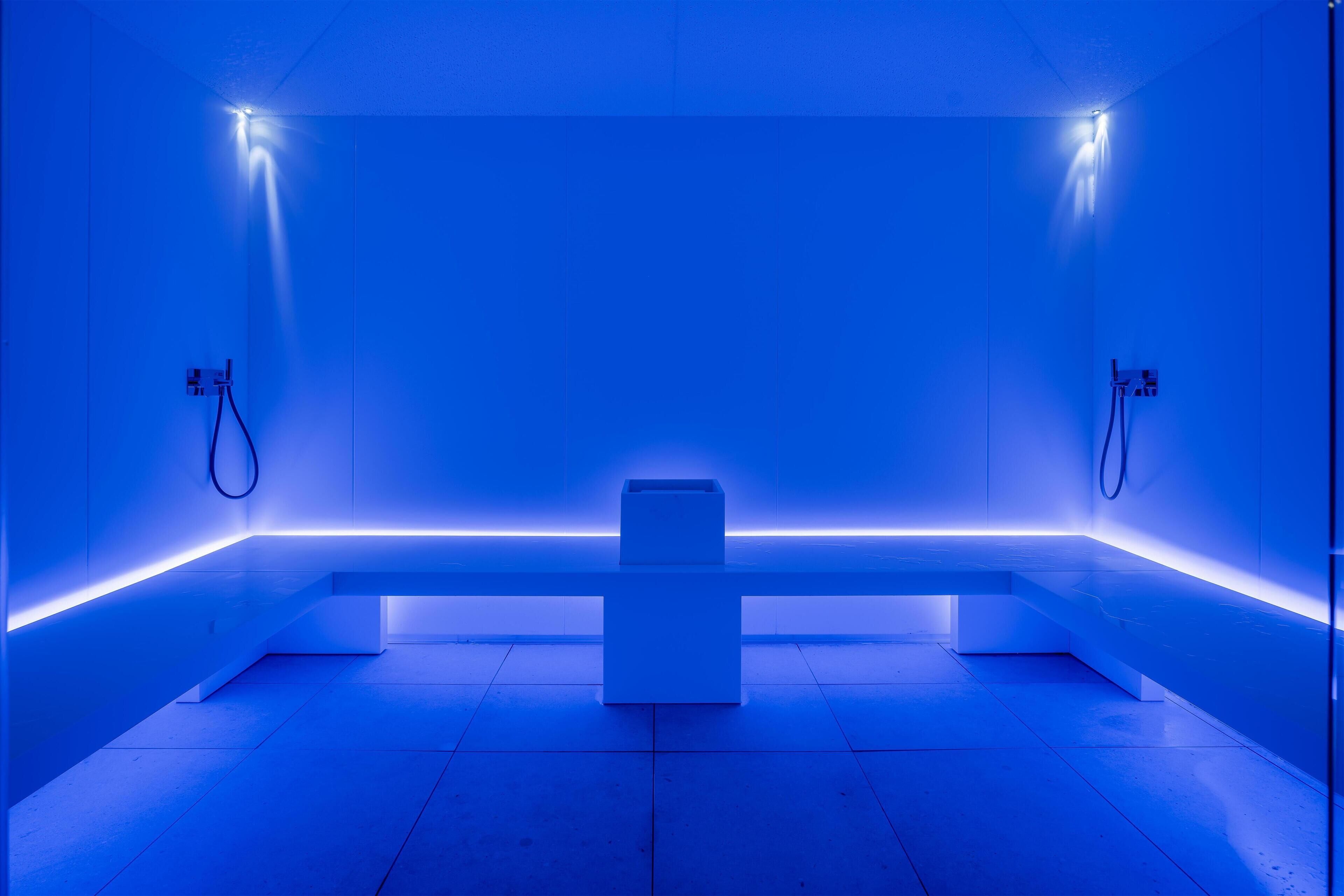 A modern, minimalist bathroom with a blue hue, featuring a central bench and two towel rings on the walls.