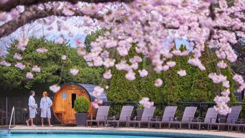 A pool area with lounge chairs, a wooden cabin, and a backdrop of trees and cherry blossoms.