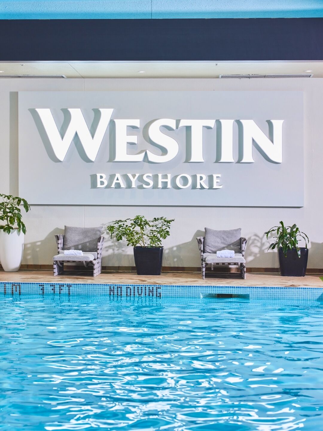 Zona junto a la piscina con sillones de descanso y plantas en macetas, un cartel que dice 'WESTIN BAYSHORE' y una piscina azul cristalina.